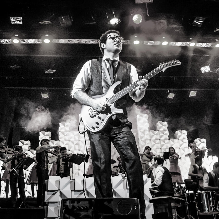 Everton Vidal Azevedo leading a guitar performance with the Camerata del Oriente ensemble behind him, black-and-white image. Una Canción Hecha Mujer. Photo by Antonio Dávila.