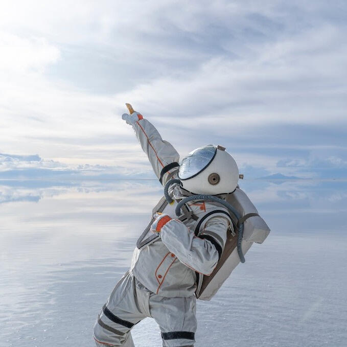 Everton Vidal Azevedo in an astronaut suit pointing toward the sky at the Salar de Uyuni. Photo by Marvin Montes.