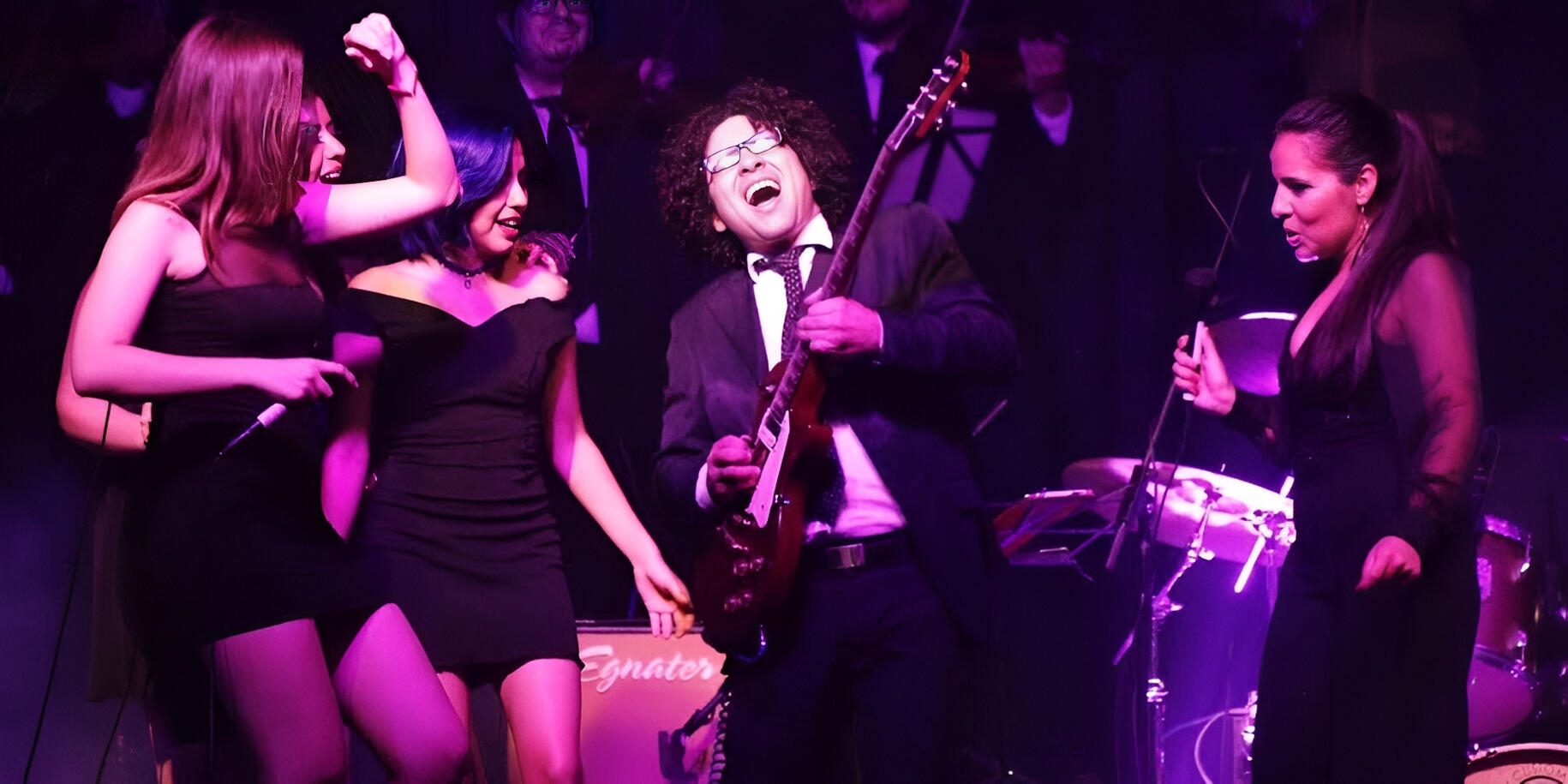 Everton Vidal performing live with the Camerata del Oriente during the Girl Power concert, sharing the stage with vocalists Jessi Luna, Yna, and Lizzett under vibrant pink and purple lights.