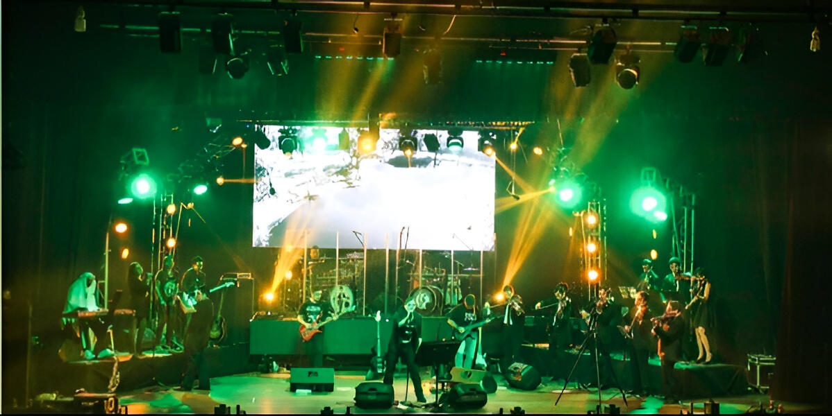 With the band Dixi and the Camerata del Oriente — supporting guitarist & arranger Everton Vidal performing as supporting guitarist and arranger with the rock band Dixi alongside the Camerata del Oriente, on a stage with orchestral musicians and dramatic red lighting.