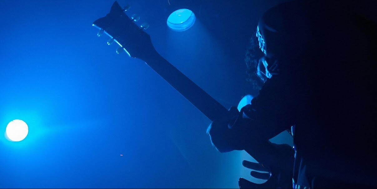 Onstage energy, live show Everton Vidal Azevedo onstage with electric guitar raised, blue backlight and atmospheric lighting.